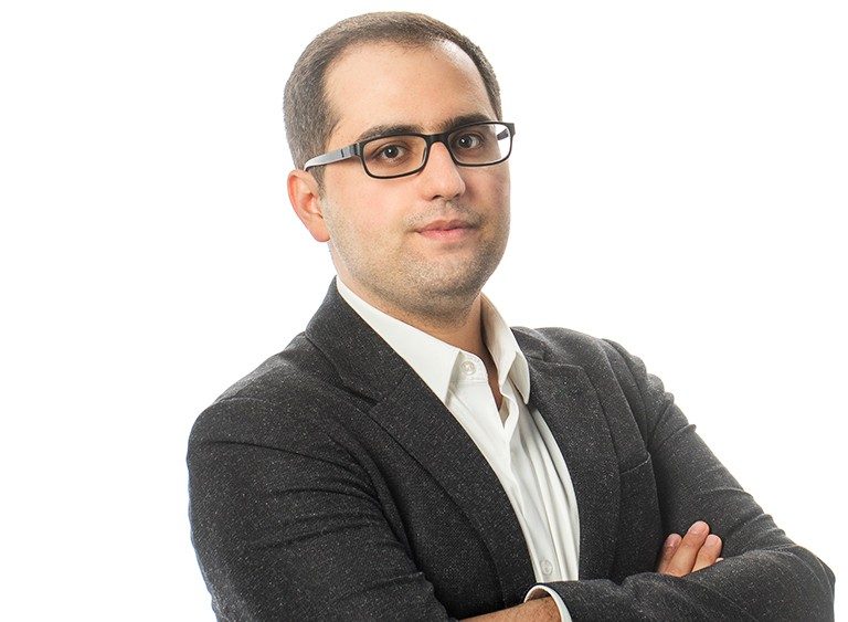 Man with very short, dark hair, dark-rimmed glasses and a brown jacket.