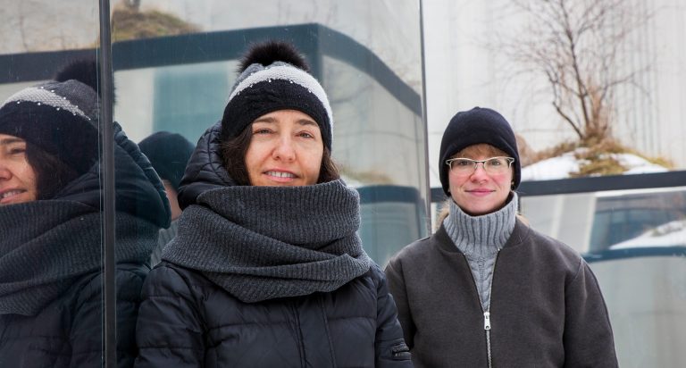 Two women standing side-by-side, both wearing winter hats and jackets.