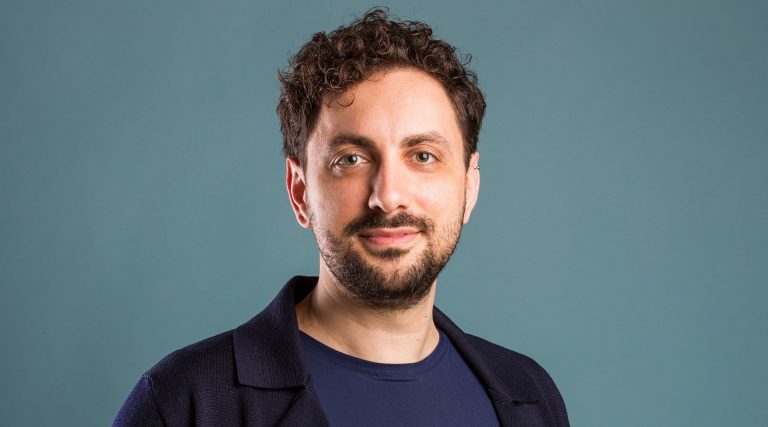 A smiling man with short, dark curly hair, a beard, an earring, a blue T-Shirt and a black jacket.