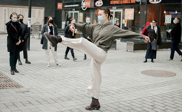 A young woman raising her leg in a dance move on a city street with people watching or walking past.