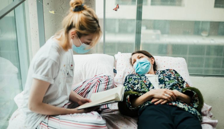 A young woman wearing a mask and sitting on bed with windows in the background, reads to an older woman lying on the bed, also in a mask.