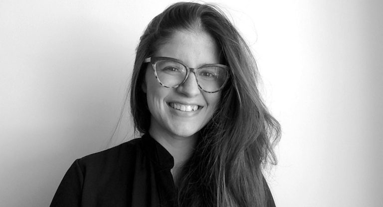Black and white photo of a young smiling woman with long, dark, straight hair, glasses and a black shirt.