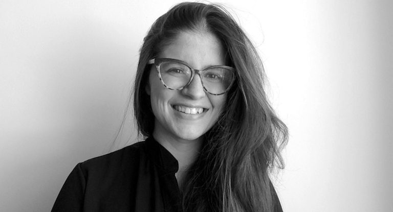 Black and white photo of a young smiling woman with long, dark, straight hair, glasses and a black shirt.