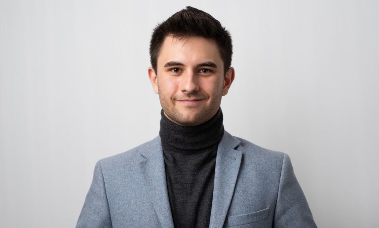 Colour head-and-shoulders photo of a young smiling man with short, dark hair, wearing a dark turtleneck under a pale blue blazer.