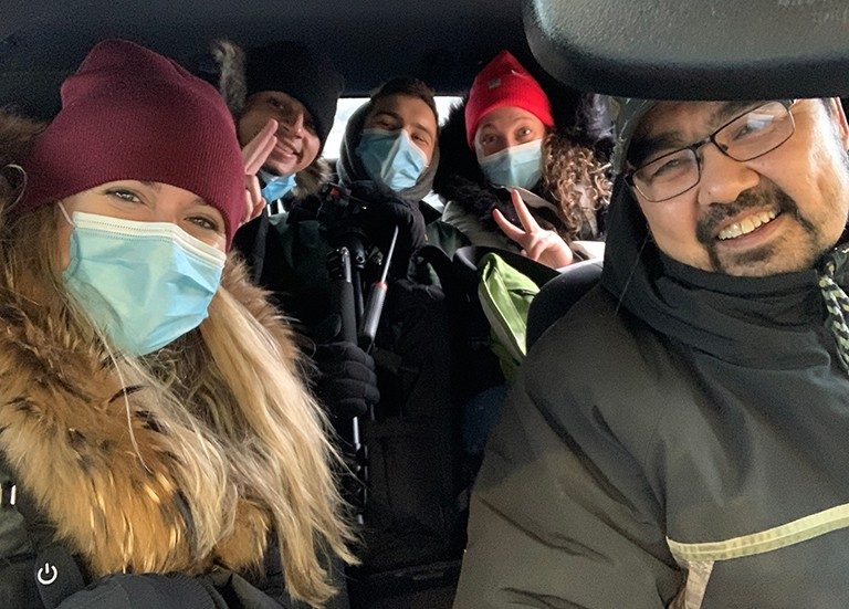 Five people sitting in a car and smiling for the camera, with three of the people wearing face masks.