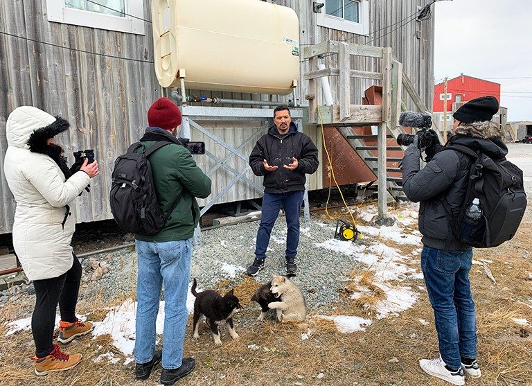 Three young people with cameras interviewing a man while three young puppies play in the foreground.
