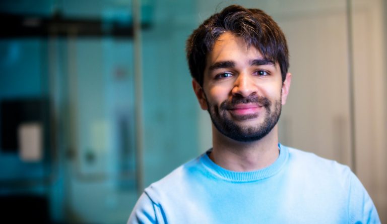 Young, smiling man with short dark hair, a beard and a light blue top.