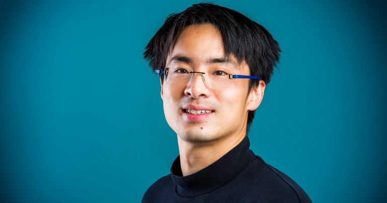 Young, smiling, Asian man with glasses, short dark hair and a black turtle-neck top.