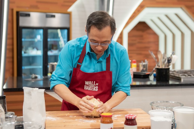 Asian man with short, dark hair, glasses, a blue shirt and a red apron that says "Wall of Bakers" kneading dough on a wooden cutting board