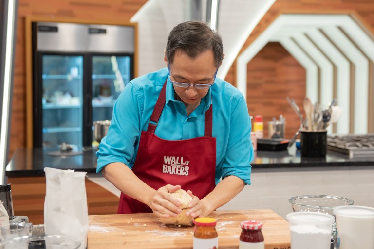 Jang Kwon | Food Network Canada Asian man with short, dark hair, glasses, a blue shirt and a red apron that says "Wall of Bakers" kneading dough on a wooden cutting board