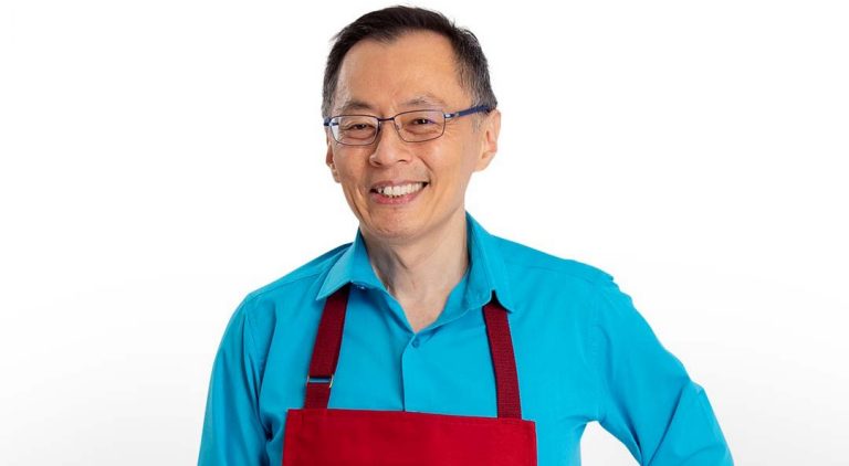Smiling older Asian man with short, dark hair, glasses, a blue shirt and a red apron that says "Wall of Bakers."