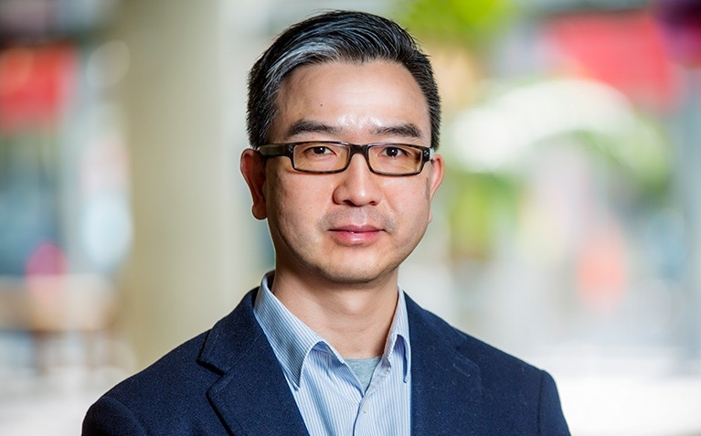 A slightly smiling Asian man with dark-rimmed glasses, short hair, a blue shirt and blue blazer.