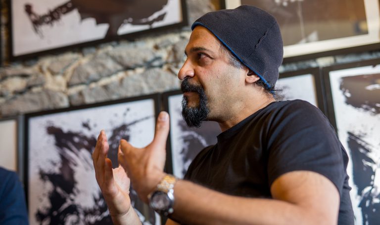 Man with a dark beard, black T-Shirt, a black beanie, talking to someone off camera.