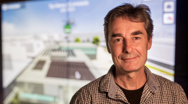 Smiling older man with short hair and a checkered shirt, standing in front of a video game.