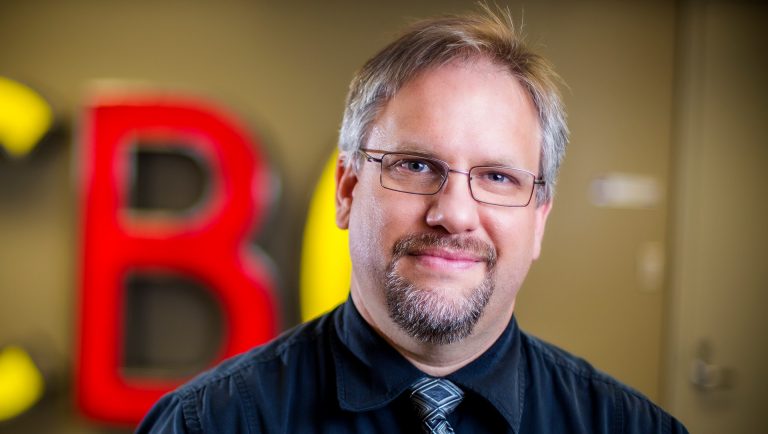 Smiling middle-aged man with short blonde-grey hair, with a beard, wearing glasses and a black dress shirt and tie.