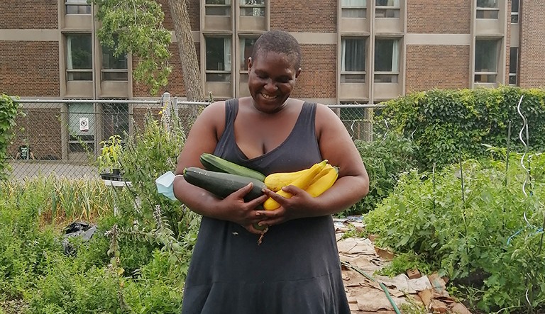 Collective member of The People's Potato, Sosa. Smiling Black woman in a black, sleeveless dress, holding courgettes in a community garden.