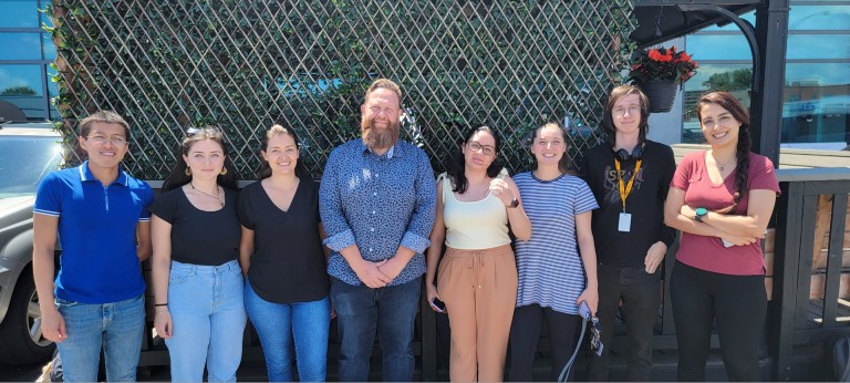 Group photo of 8 people standing outside in front of green lattice wooden structure