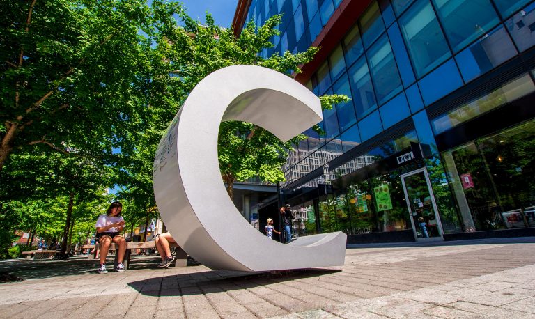 Downtown urban area with trees and people and in the foreground a large sculpture of the letter "C".