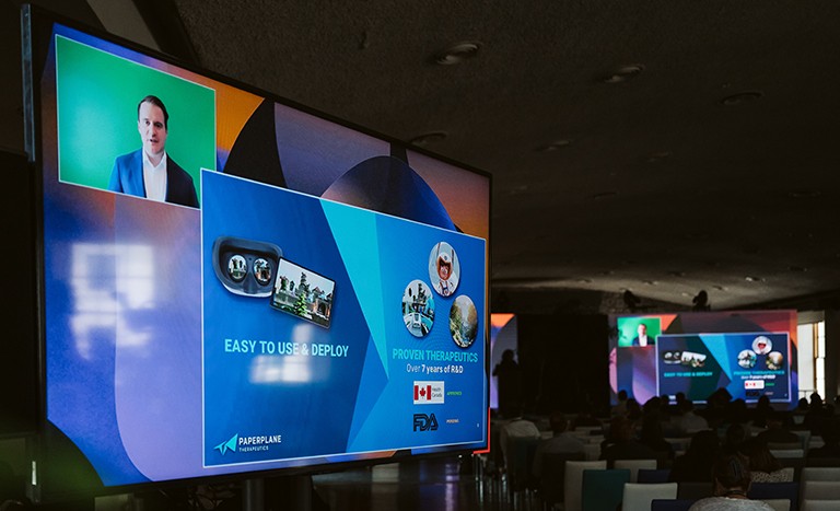 Image of a auditorium with an audience and in the forefront a large television screen with a powerpoint presentation.