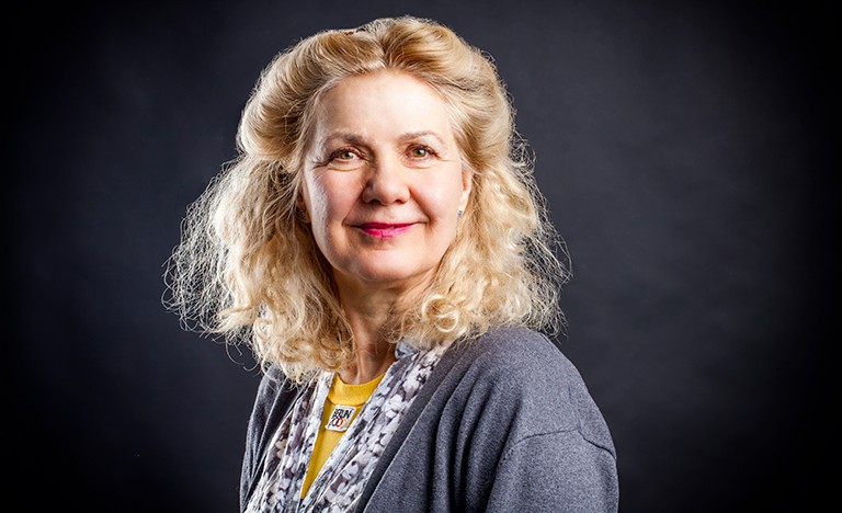 Smiling woman with curly, long hair pulled back from her face, wearing a printed shirt and a grey cardigan