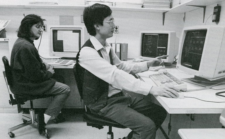 A black-and-white archive photo of two men sitting at a desk with old desktop computers.