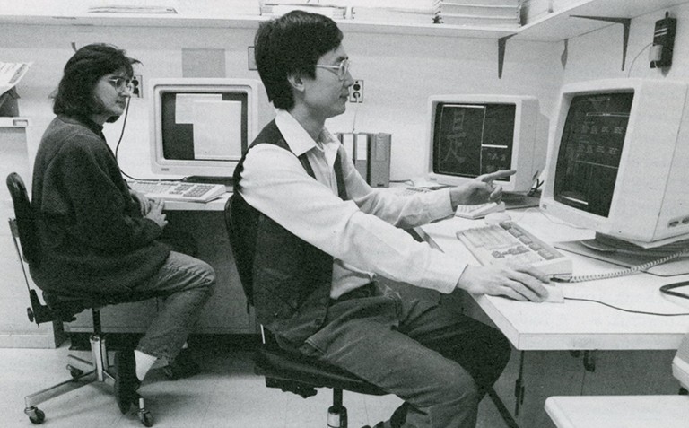 A research assistant, Christine Nadal, sits with analyst William Wong in 1990. A black-and-white archive photo of two men sitting at a desk with old desktop computers.