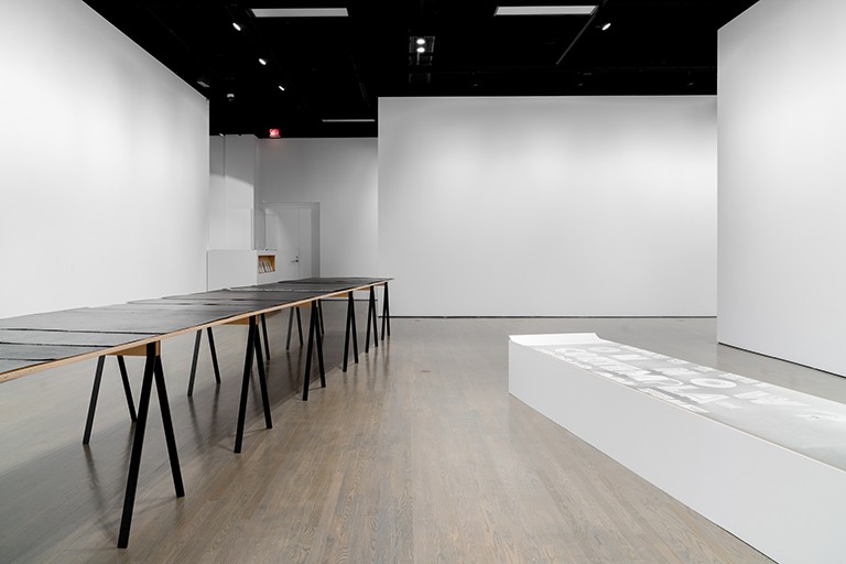 View of works by Thérèse Mastroiacovo. Curator Michèle Thériault. | Photo Jean-Michael Seminaro Art exhibit: tables with black legs and with dark paintings on top, in a room with spare white walls