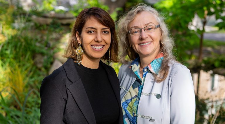 Two smiling women, one younger and one older, standing together smiling at the camera in an outdoor setting.