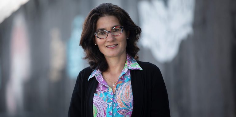 Smiling woman with shoulder-length dark hair, red-rimmed glasses and wearing a blue top and red necklace.