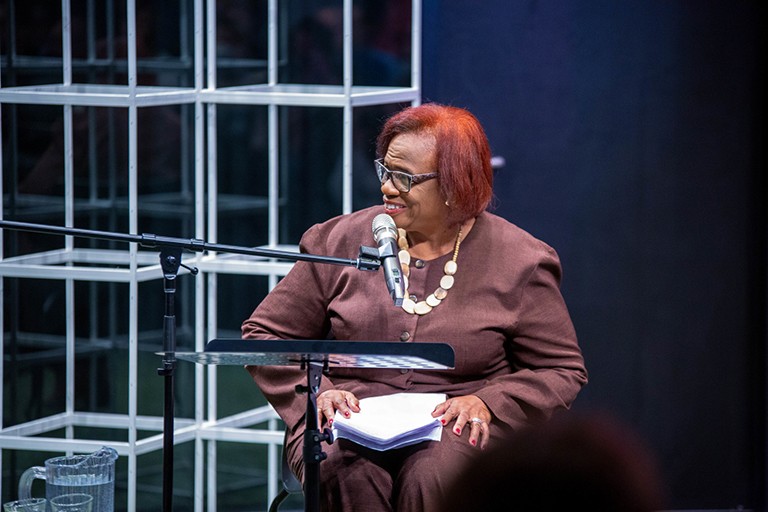 A Black woman sitting on a chair and smiling to the left, with papers in her lap.