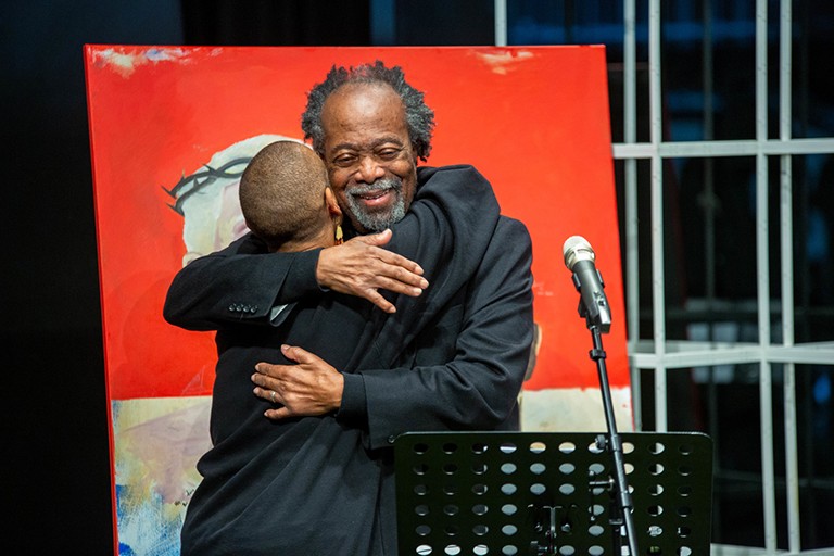 From left: Angélique Willkie with Rodney John who shared his experiences of racism at SGWU. A Black man hugging a Black woman with a microphone in the foreground and a painting in the background.