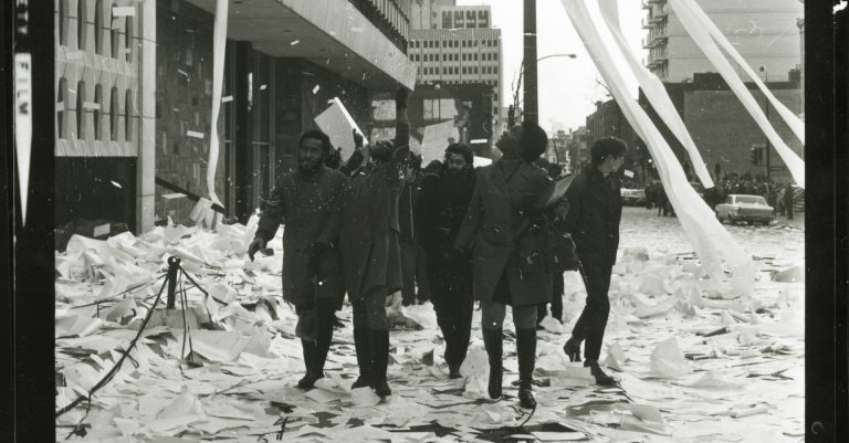 Archive black and white image of students walking through reams of paper on the pavement.