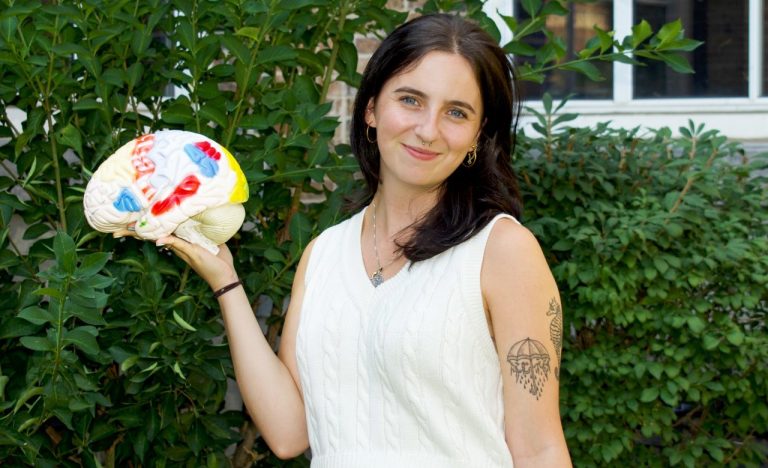 Smiling young woman with long dark hair standing in front of a hedge, wearing a white sleeveless knit top and holding a plastic model of a brain.