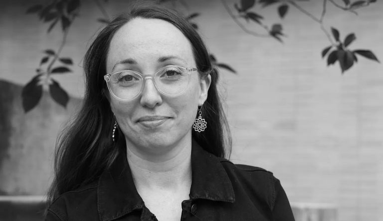 Black and white image of a smiling woman with long, dark hair, wearing glasses and a black shirt.