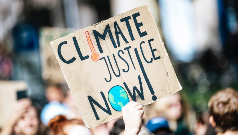 Hand holding up a sign saying, "Climate justice now!" with a crowd in the background.