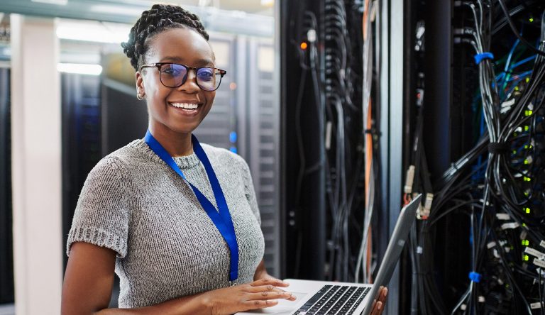 Smiling woman with braided dark hair and wearing glasses standing with a laptop in front of servers.