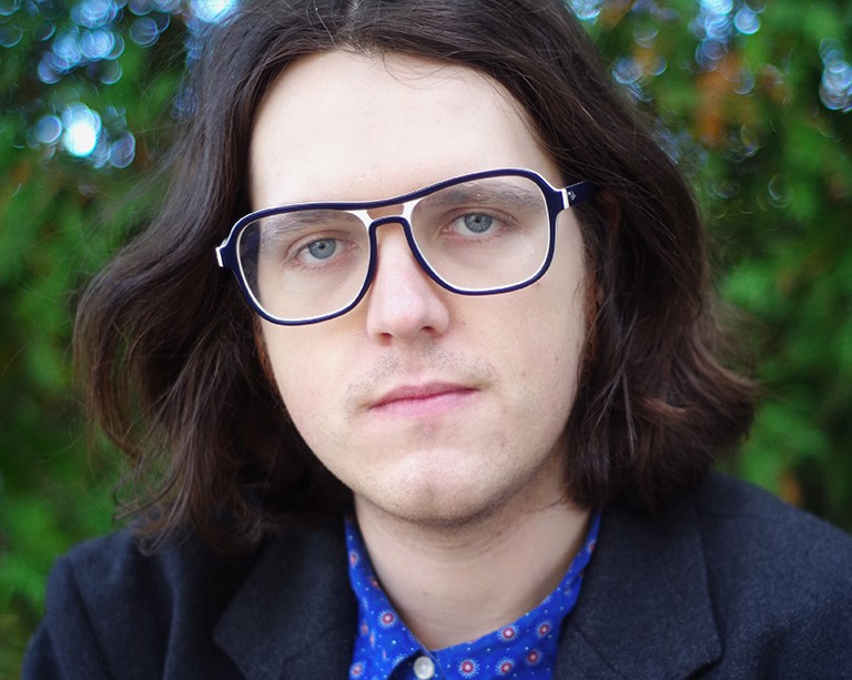 Young man with dark-rimmed glasses, shoulder-length dark hair and blue eyes.