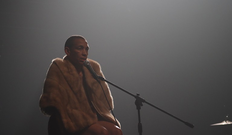 Women sitting on a stool, wearing a beige fur coat, and speaking into a microphone