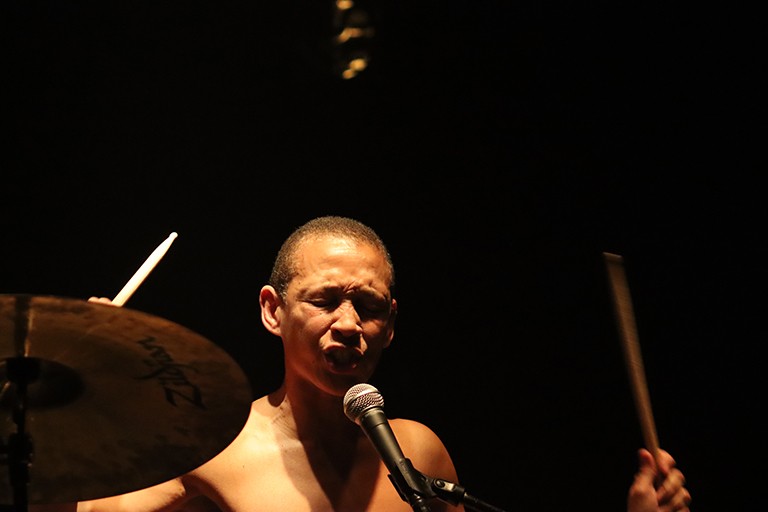 A woman with her eyes closed and speaking into a microphone while she plays drums.