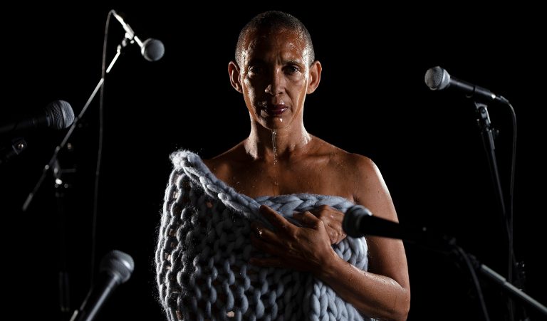 A woman staring directly at the camera, with shaved dark hair and water dripping down her face, standing with a grey knitted material draped around her, and microphones placed to the sides and turned towards her.