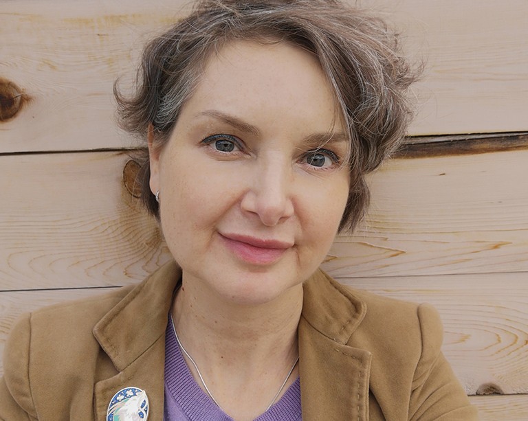 Smiling woman with greying, dark-blonde hair, standing in front of a wooden wall and wearing a beige blazer and purple sweater.