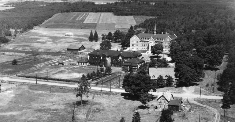 Black and white aerial photo of school buildings in a large, cleared area of fields in the countryside.