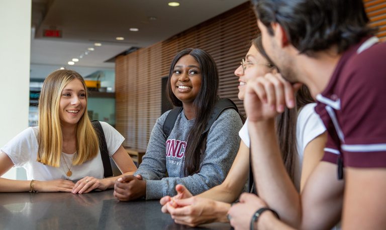 A group of three young smiling women and one man, sitting at a table in an atrium-style interior and talking together.