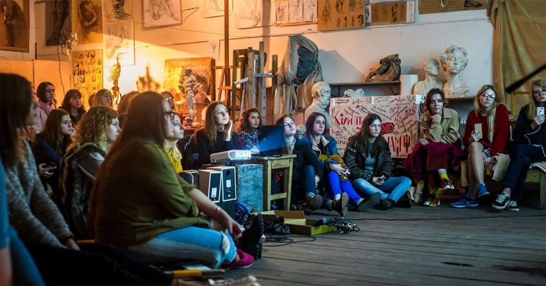A group of young women sitting in a cluttered art space and watching a film being played from a projector.