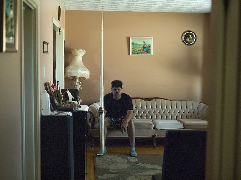 A young man in a black T-Shirt sitting on a sofa in a salon and holding a white tapered object that is nearly touching the ceiling.