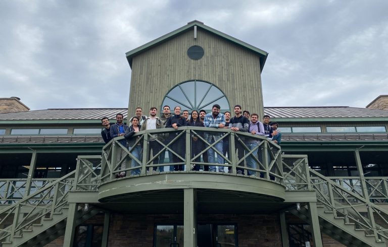 17 young adults standing on balcony of two-storey green building, smiling down at the camera.