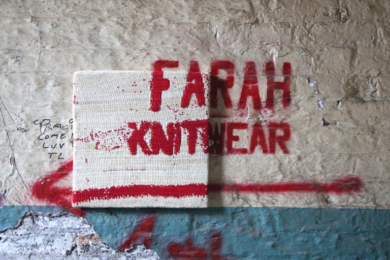 Piece of knitted light fabric on a small canvas on wall, with "Farah Knitwear" in red spray-painted partially on canvas and partially on wall
