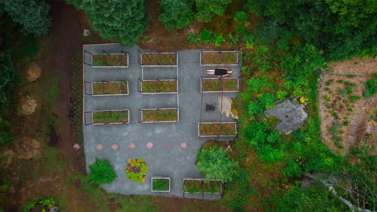 Overhead shot of woman in black dress lying on her back on the gravel between 9 small vegetable gardens