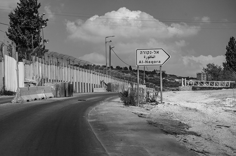 Black and white image of a road with desert on one side and a wall on the other, with a road sign in Hebrew.