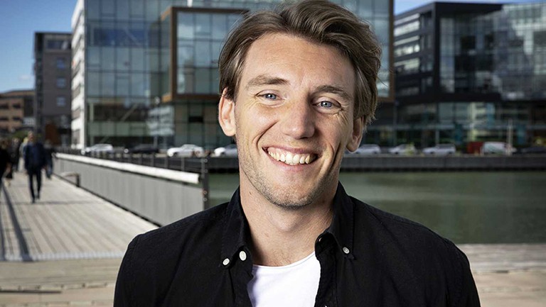 Young smiling man with short, blonde hair, wearing a white T-Shirt and a black collard shirt.